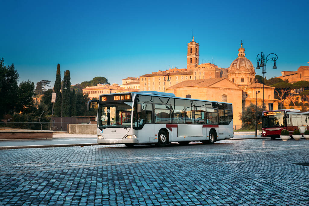 Rome, Italy. Public Bus Driving In Via Dei Fori Imperiali Street In Sunny Summer Morning Near Santi Luca E Martina Church.