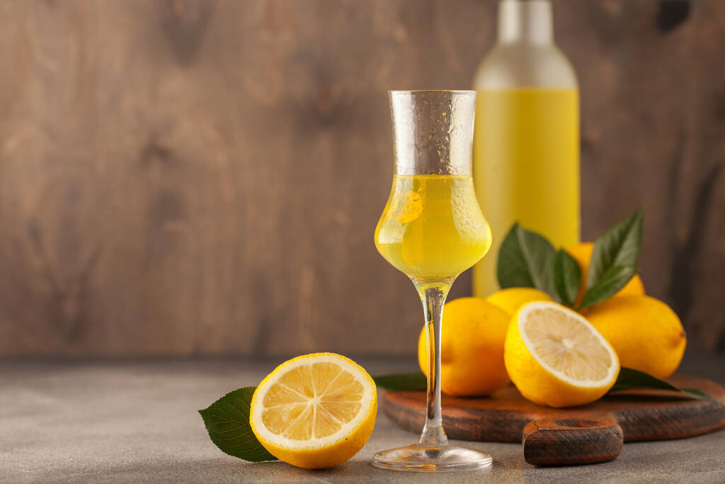 Limoncello in glass, sweet Italian lemon liqueur, traditional strong alcoholic drink and lemons on the grey background.
