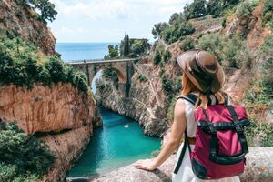 Plan podrózy po Wybrzeżu Amalfi. A girl in a cowboy hat with a backpack enjoys an incredibly beautiful view of a stone bridge over the gorge Fiordo di Furore. Little bay with paradise beach. Back view. Amalfi, Italy